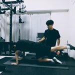 パーソナルジムでトレーナーの補助を受けながら、四つ這いで片脚を上げ、姿勢を正確にコントロールするバランス調整トレーニング。