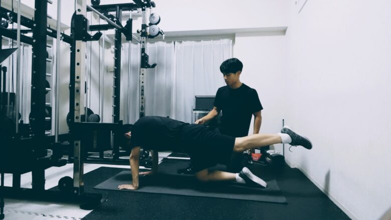 パーソナルジムでトレーナーの補助を受けながら、四つ這いで片脚を上げ、姿勢を正確にコントロールするバランス調整トレーニング。