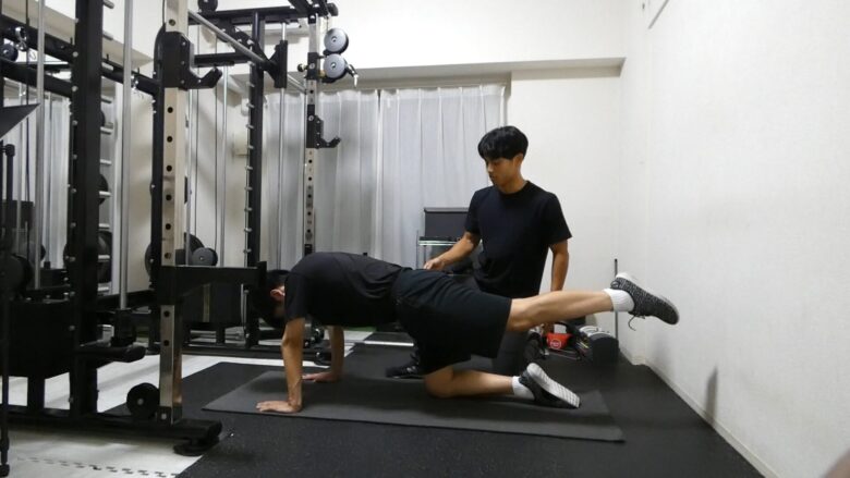 金山にあるパーソナルジムで、30代の男性がトレーナーの指導のもと、ドンキーキックに挑戦している様子。体を引き締めるためのトレーニングです。
