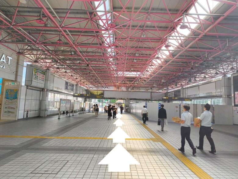 金山総合駅の連絡通路。ただたんパーソナルジム金山へ向かうための北口・南口方面への直進ルート。天井が赤い格子状の通路に、進行方向を示す白い矢印が表示されている。