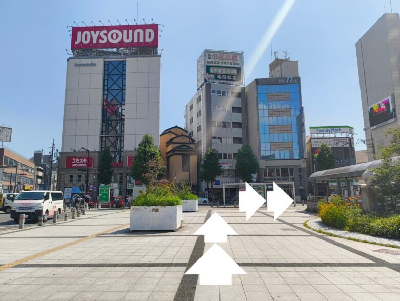 金山駅の出口付近にある開けた空間。ジョイサウンドのビルを正面に見て、右側の通路へ進むルートを白い矢印で表示している。ただたんパーソナルジム金山へ向かうための分岐点。