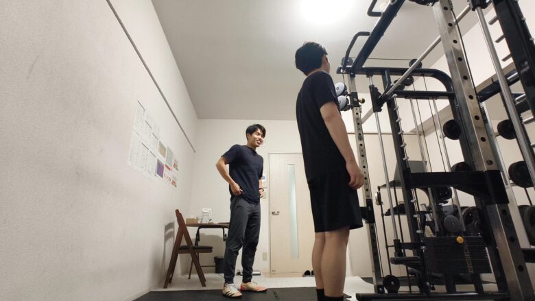 明るい照明のジム内で、笑顔で会話を交わす男性トレーナーと利用者のリラックスした様子。