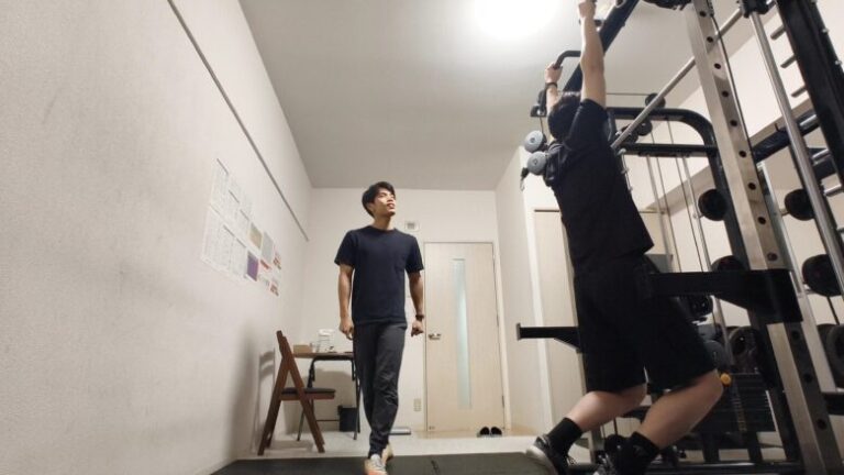スミスマシンのマルチバーを使い、懸垂を行う利用者と、その動作を真剣な表情で見守るパーソナルトレーナー。