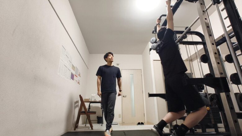 スミスマシンのマルチバーを使い、懸垂を行う利用者と、その動作を真剣な表情で見守るパーソナルトレーナー。